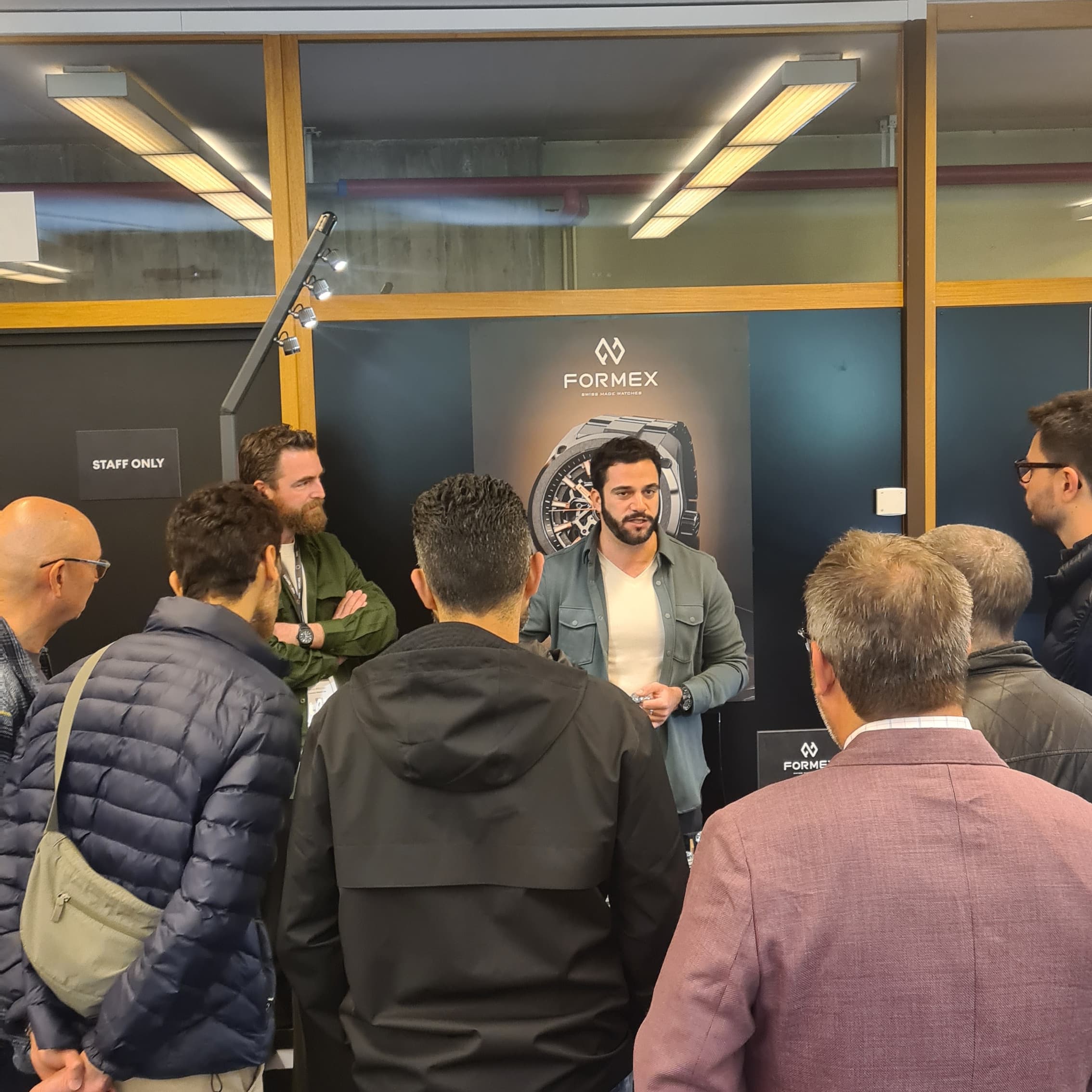group of men listening to another man talking about Formes watches in frontof stand with dark background and poster of Formex on the wall