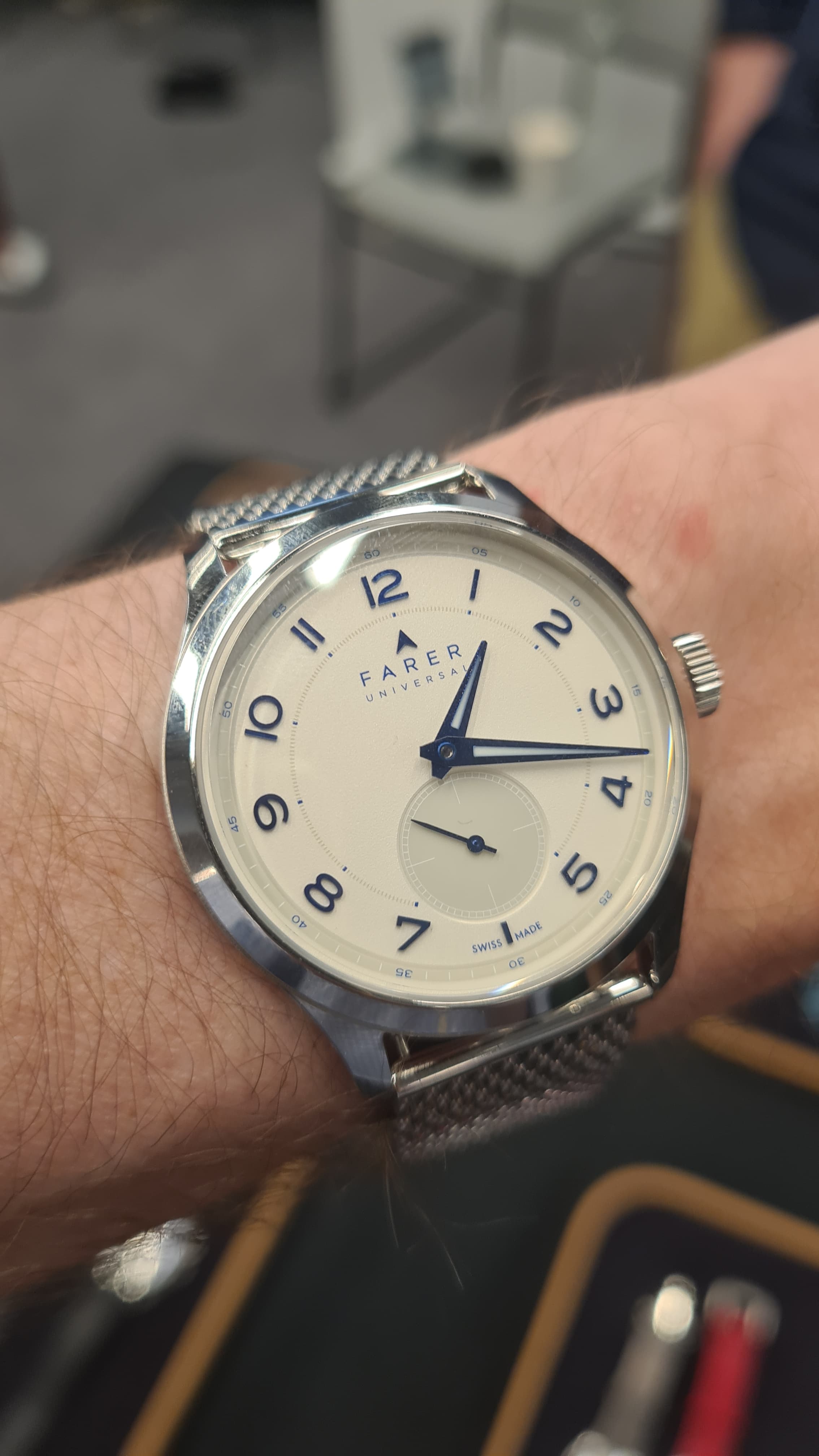 A white Farer wristwatch with dark blue numerals, hands with a grey subseconds dial on a steel mesh strap