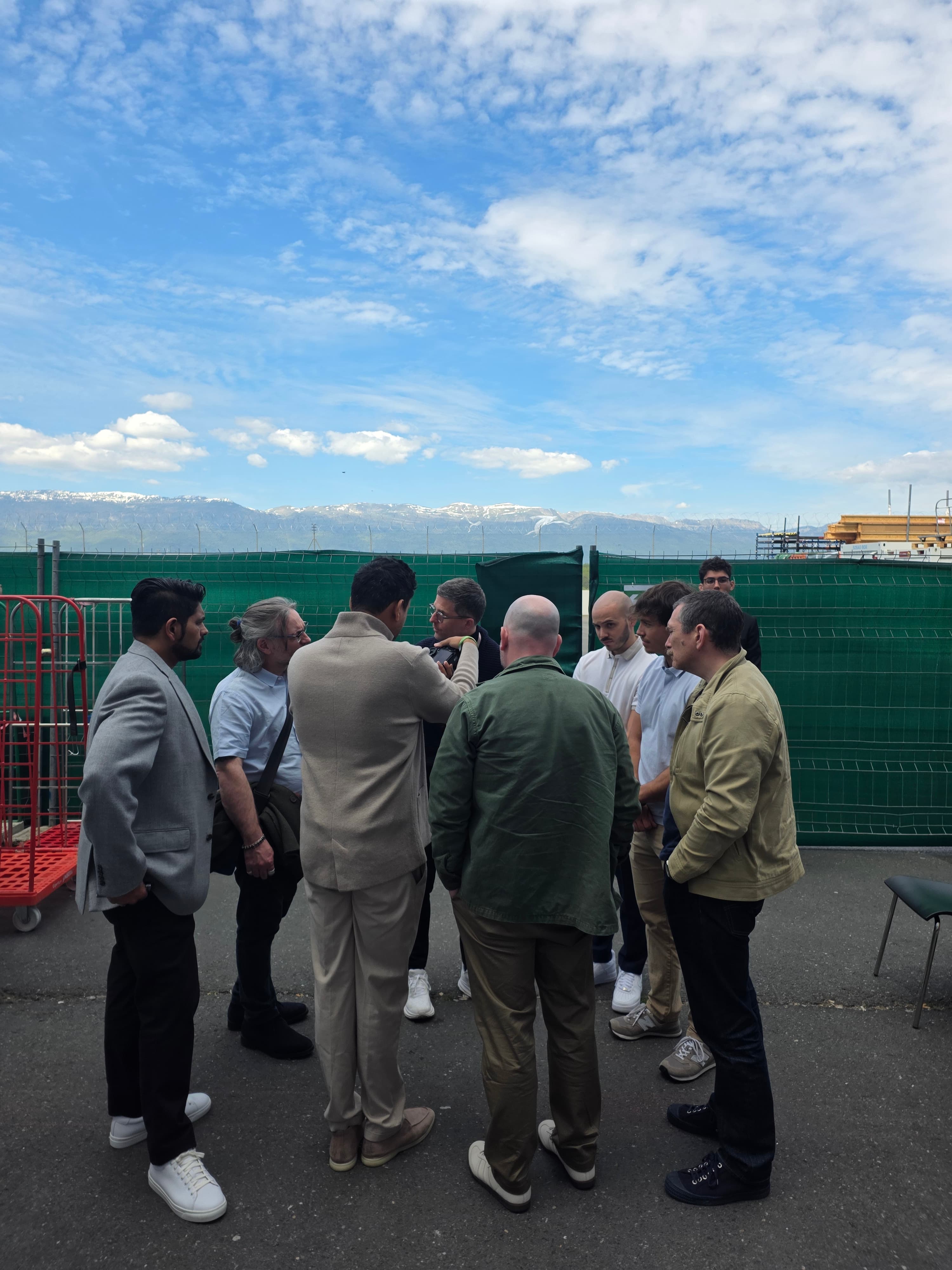 a group of men watch collectors surround soeone taking a photo of a watch with blue skies and mountains in the background