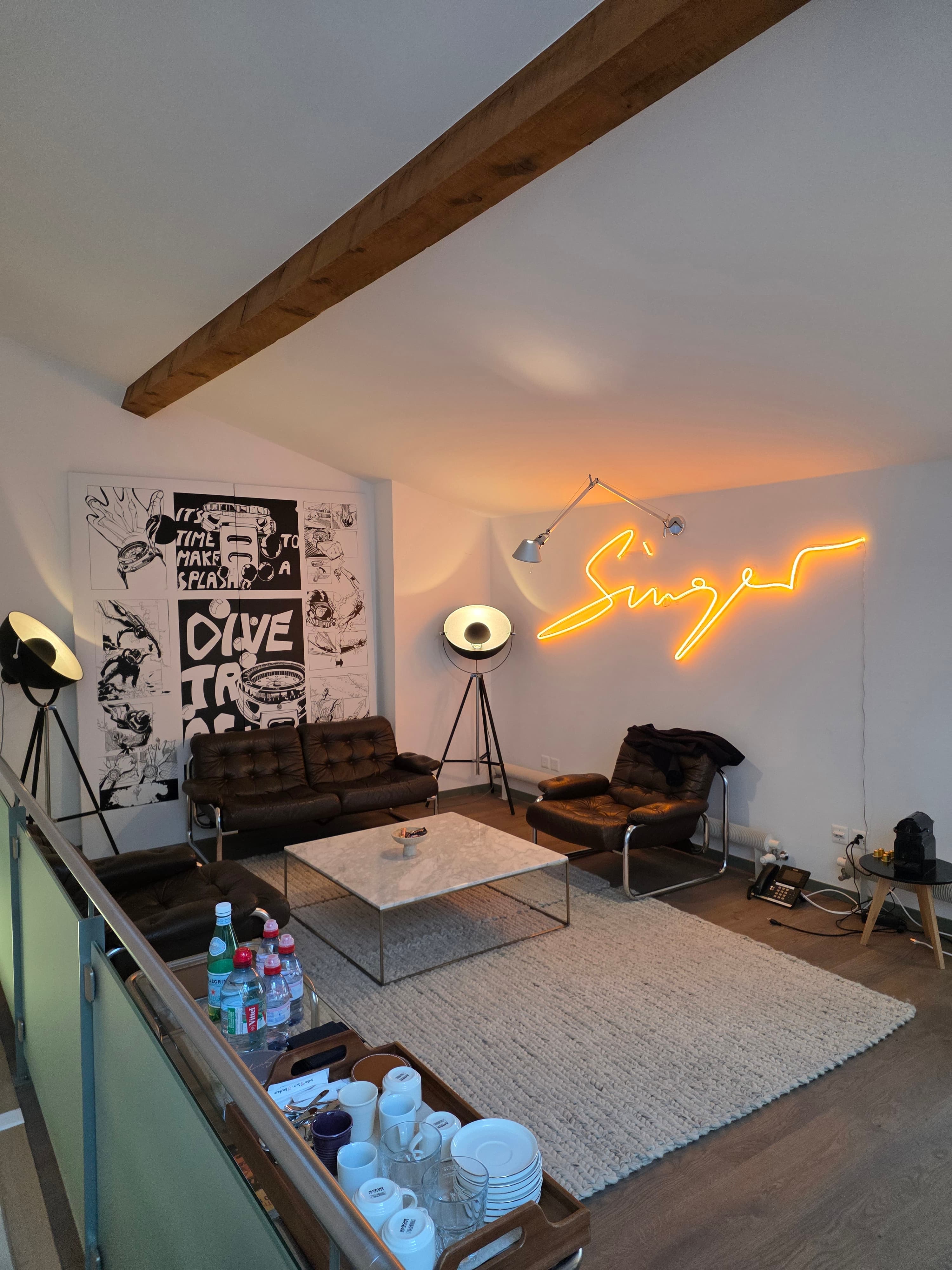 awhite painted room with a pitched ceiling, mid century furnitues and marble coffee table and beige rug, with neon Singer sign on the wall and other promotional artwork for Singer watches