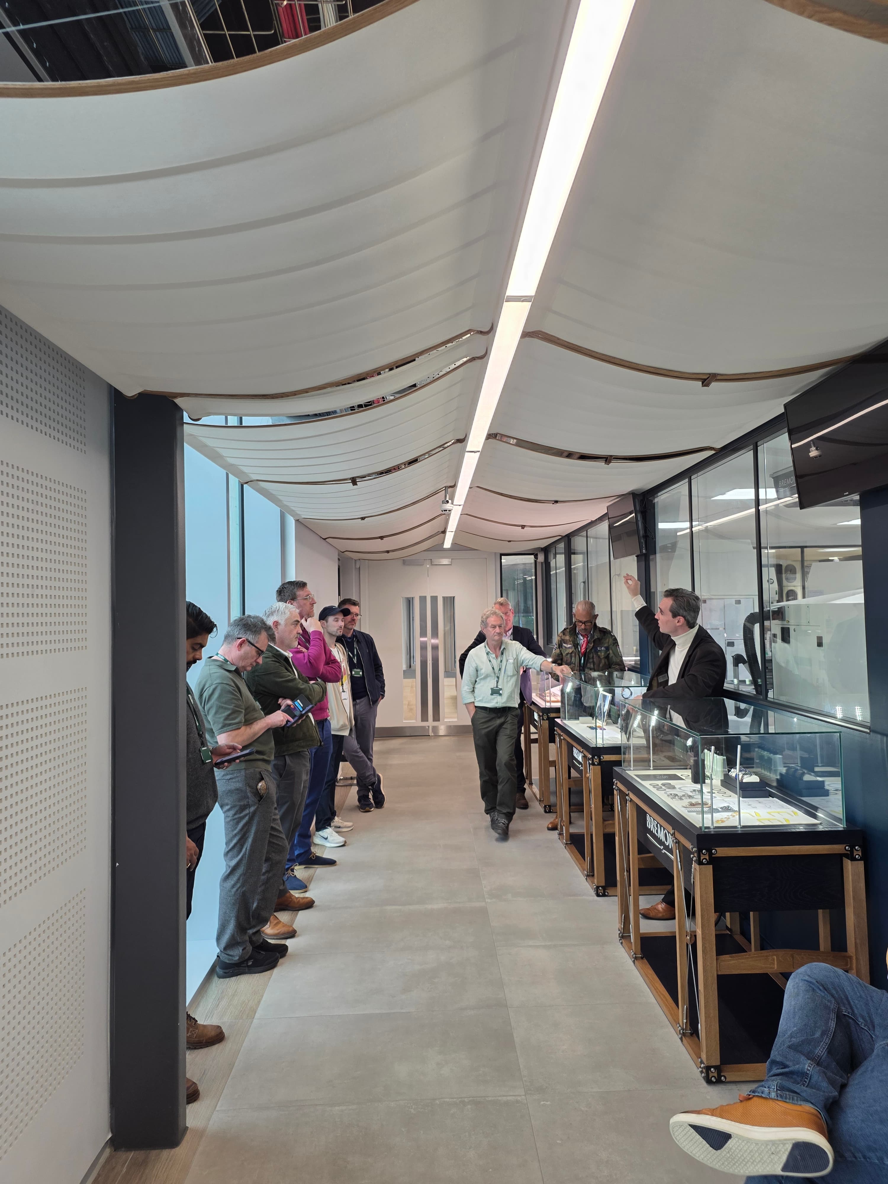 mens tanding in long grey corridor listening to talk from Bremont on their assembly process, windows on left and glass windows onto workshop on the right with strip lights on top and glass watch cabinets along the right hand side.
