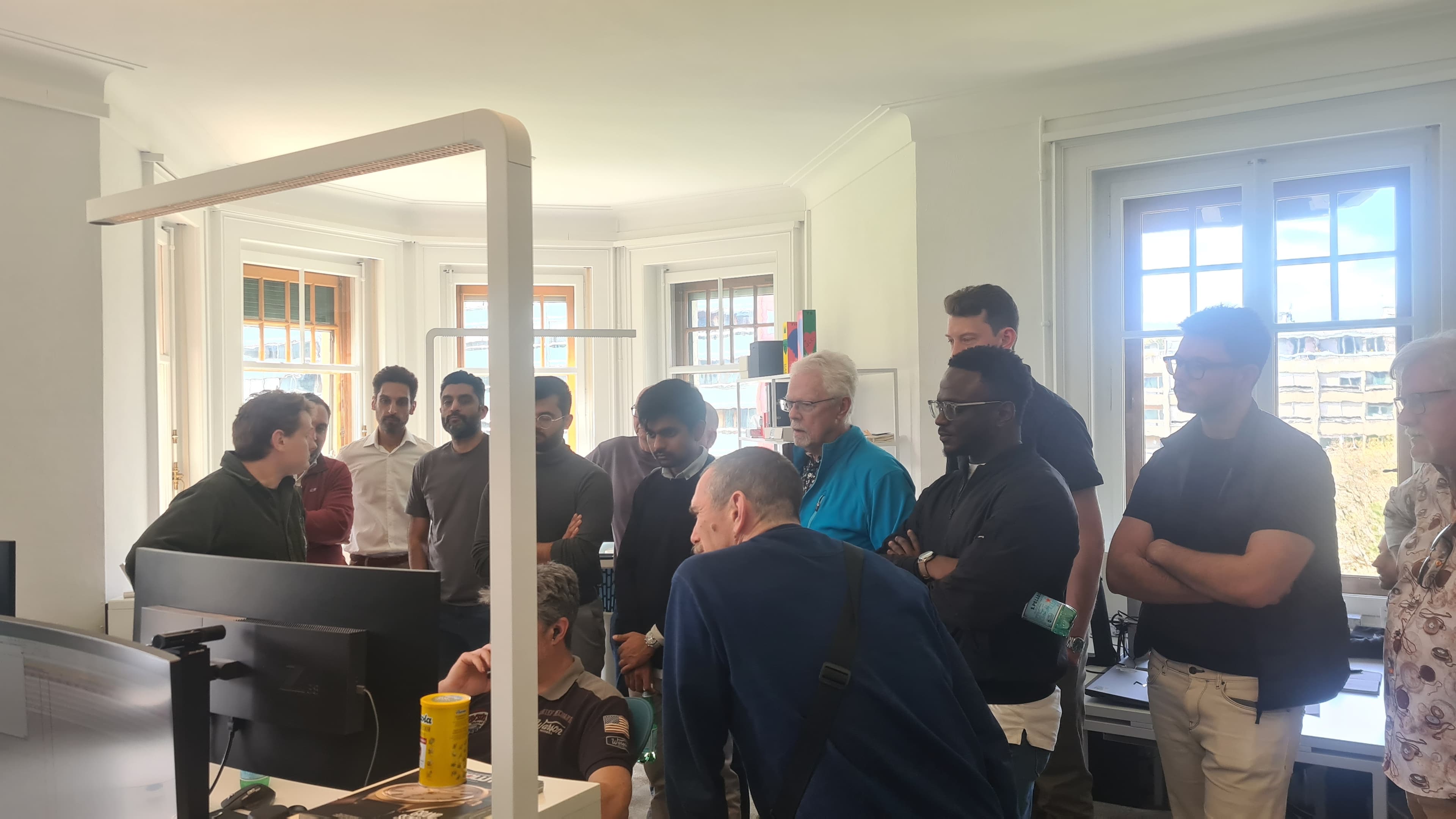 group of men standing round man sitting at desk all looking at his screen in white room with windows in the background and smart digital lighting, books on shelves in background too at M.A.D House