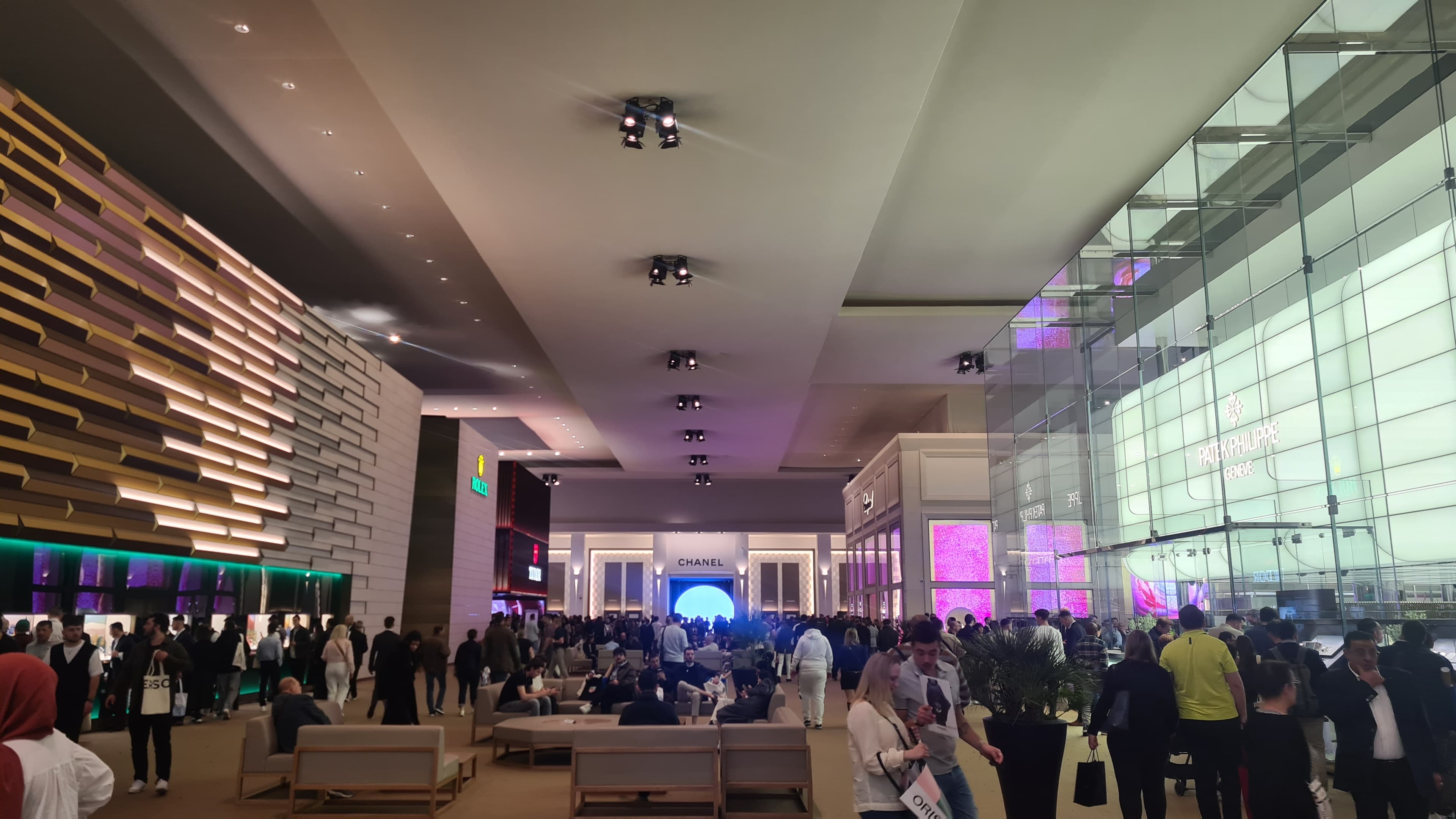 large hall of the wathes and wonders show like a mall with white ceiling and brand booths on eithe rside with glass walls, gold wars,big logos and more, full of people on the groun big crowds walking towards and away from teh viewer