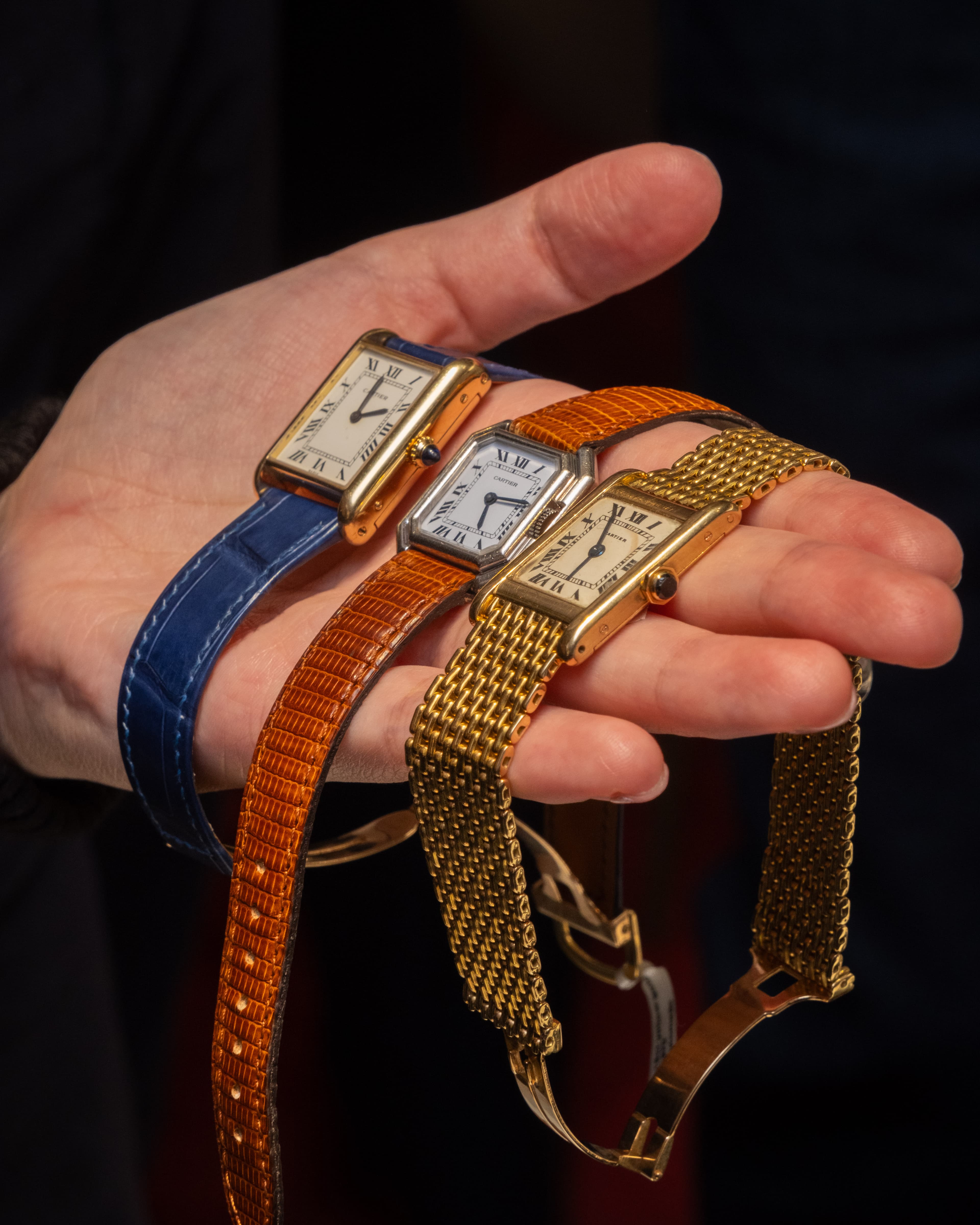 three rectangular watches being held in the palm of a hand all with white or cram faces but with different straps, one blue leather, one brown alligator leather and one gold mesh, all are Cartier at Somlo London