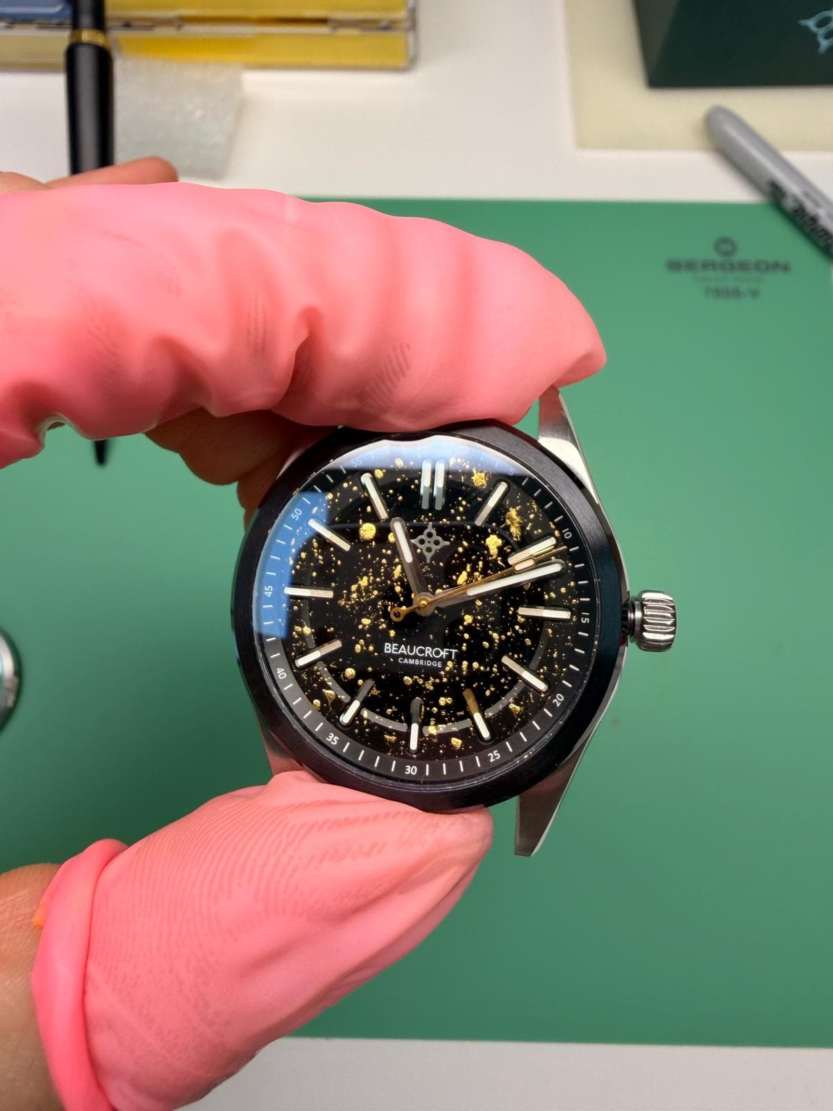a watchmaker holding a protoype of the club watch beaucrfot element in two fingers with pink finger guards the watch has a black bezel and dial with gold spots