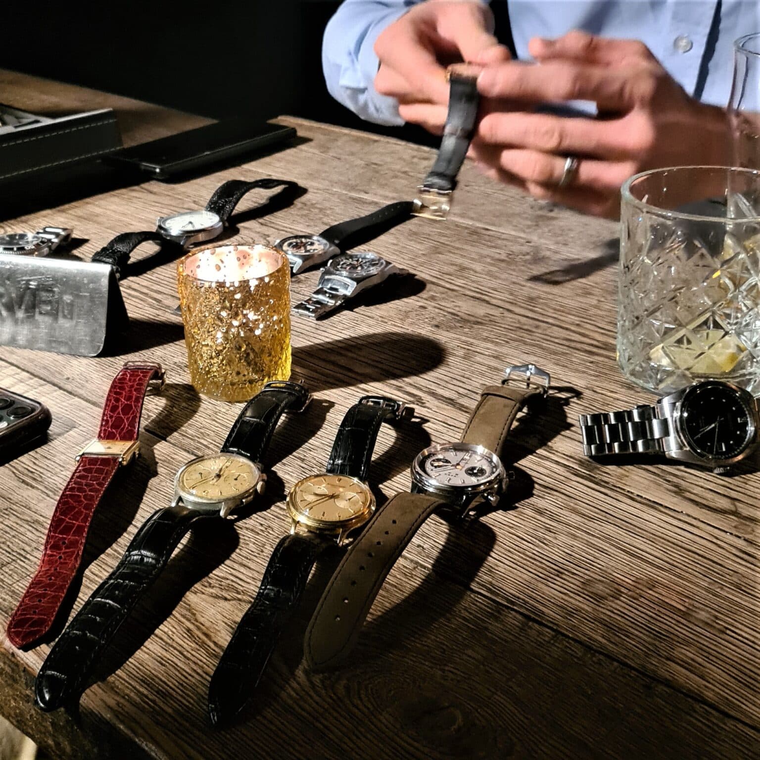 watches of different kinds arranged on a wooden table at a watch meetup with a candle and a drinks glass in the background