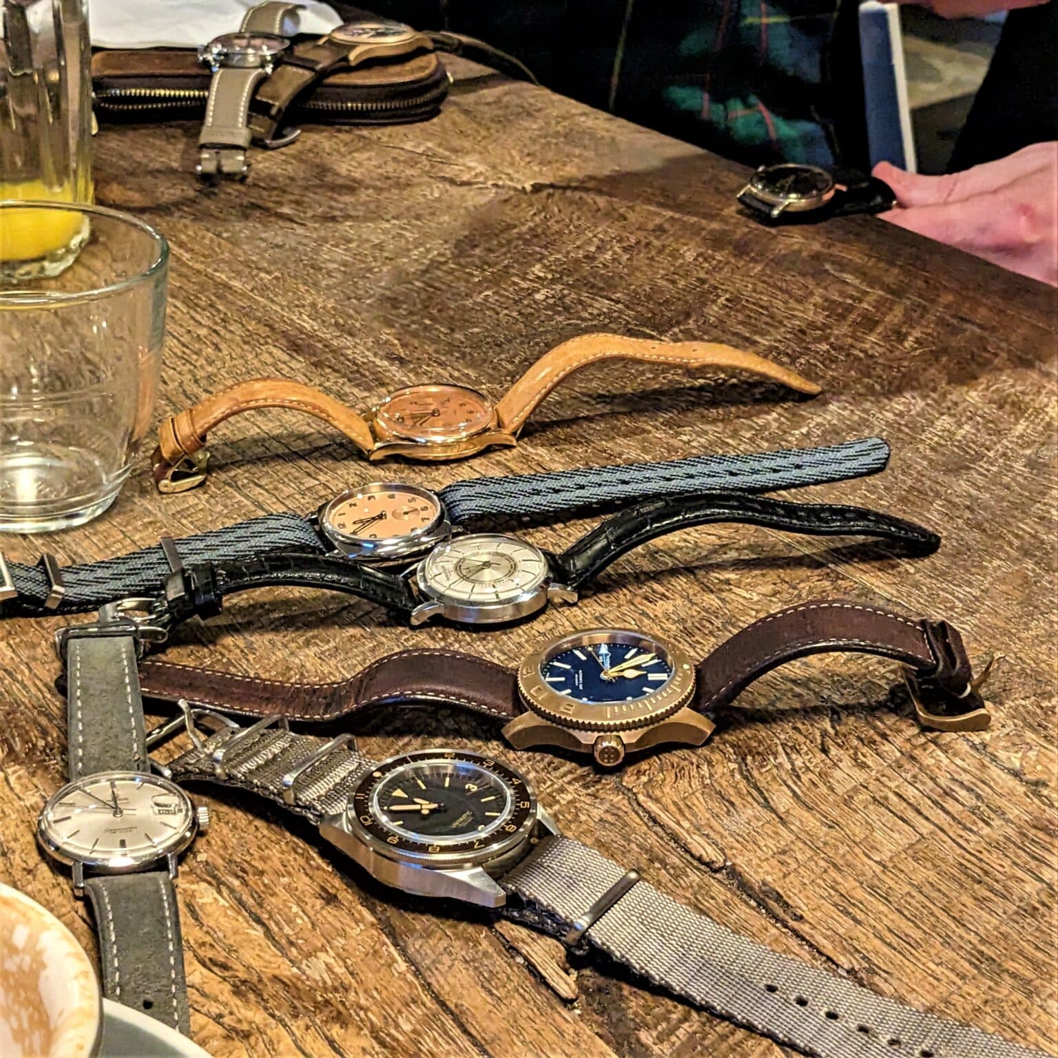 group of watches on a table at a watch meetup alongside a whiskly fglass and candle while someone looks closely at a watch in the background