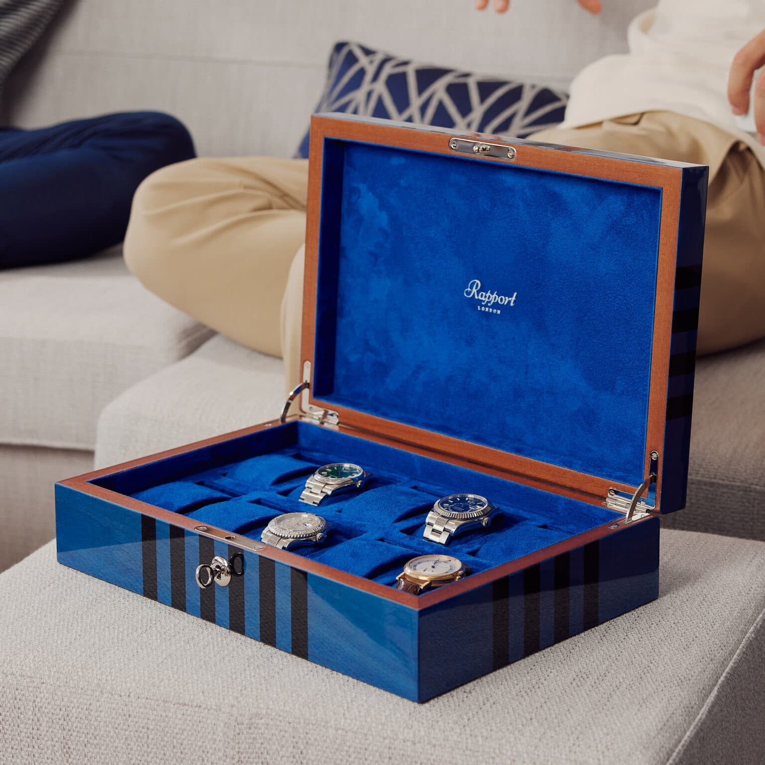 A large wooden watchbox from Rapport London with blue interior in velvet, spaces for 8 watches all in blue velvet and a blue and black lacquered wood exterior, sitting on a grey ottoman cushion