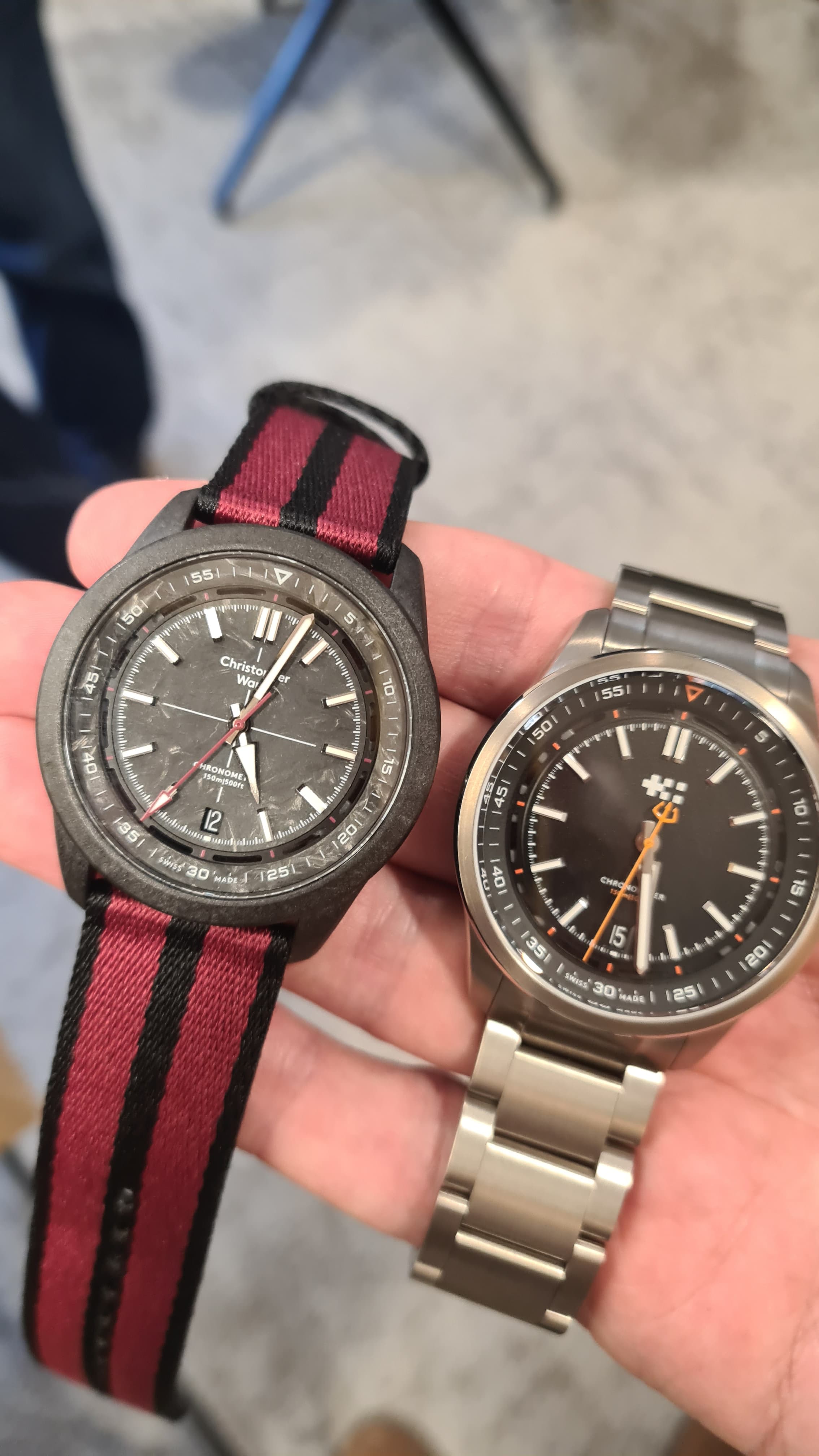two Christopher Ward C63 watches sit on the palm of a hand both with black dials white hands. the left is made of carbon fibe including the dial with fed and black fabric strap, the right is black with an orange second hand on a steel bracelet.