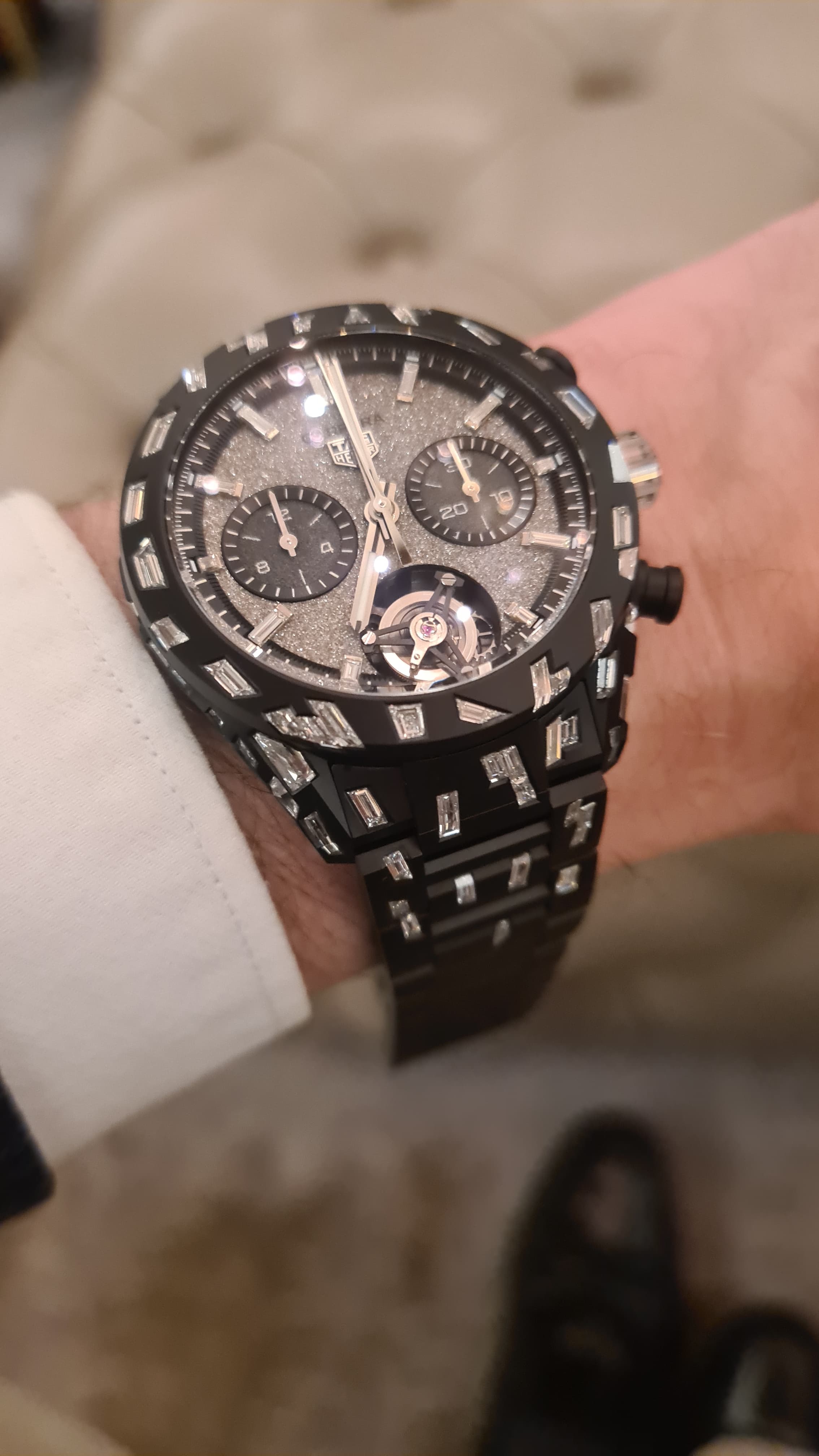 Close-up of a luxury wristwatch with a black strap, intricate dial, and silver accents, worn on a person's wrist in a formal setting.