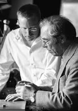 a picture of george daniels and roger smith, roger wearing a white shirt and glasses peering down at georg holding watch wearing a grey jacket, both next to a watchmakers bench