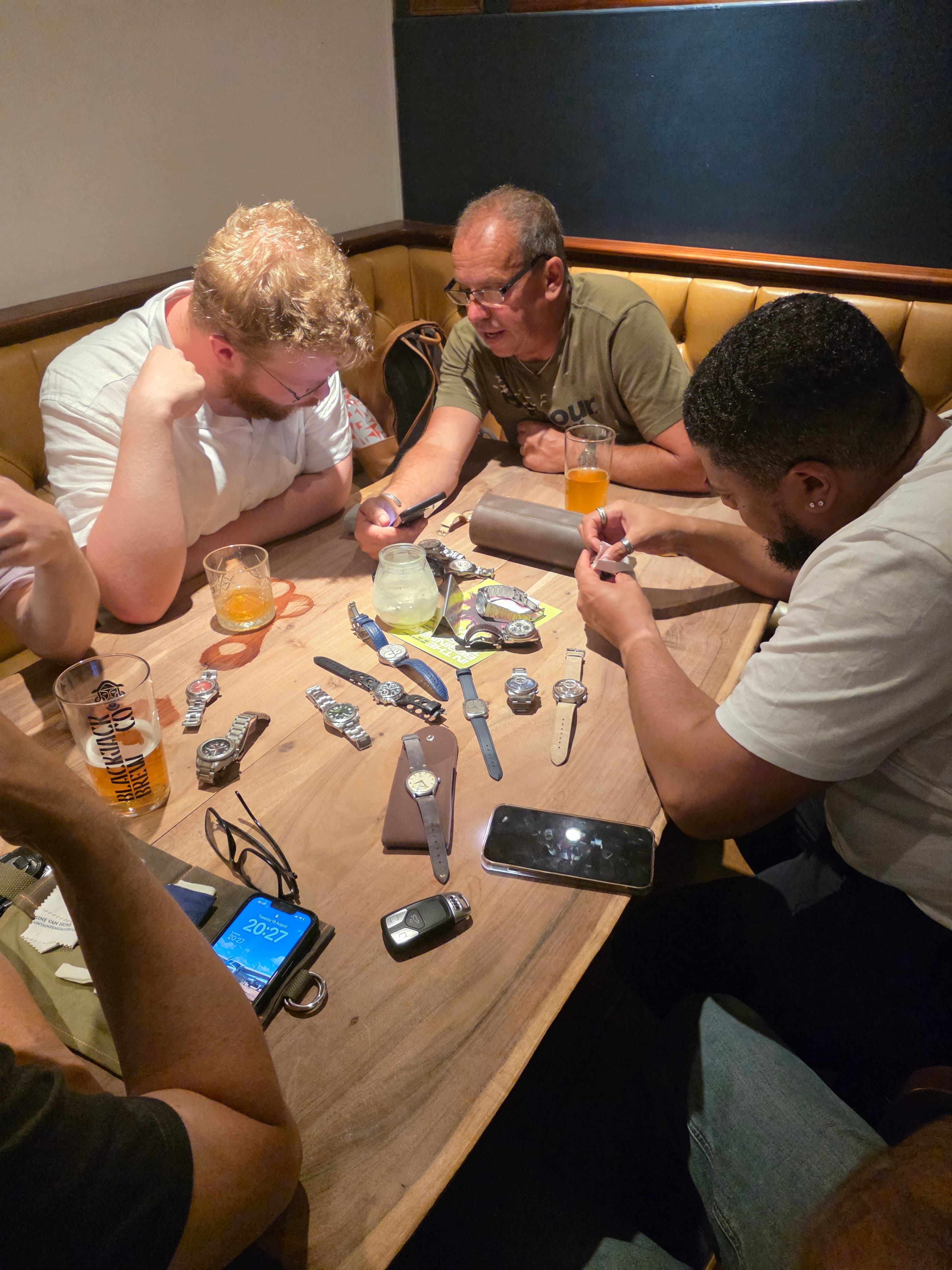five men surrouna d light wooden table with many watches on the table between them with fifferent cases bracelets and straps, the men are peeting closely at the watches there are also beers and drinks and phones on the table also, from our Manchester Watch Meetup in August 2025