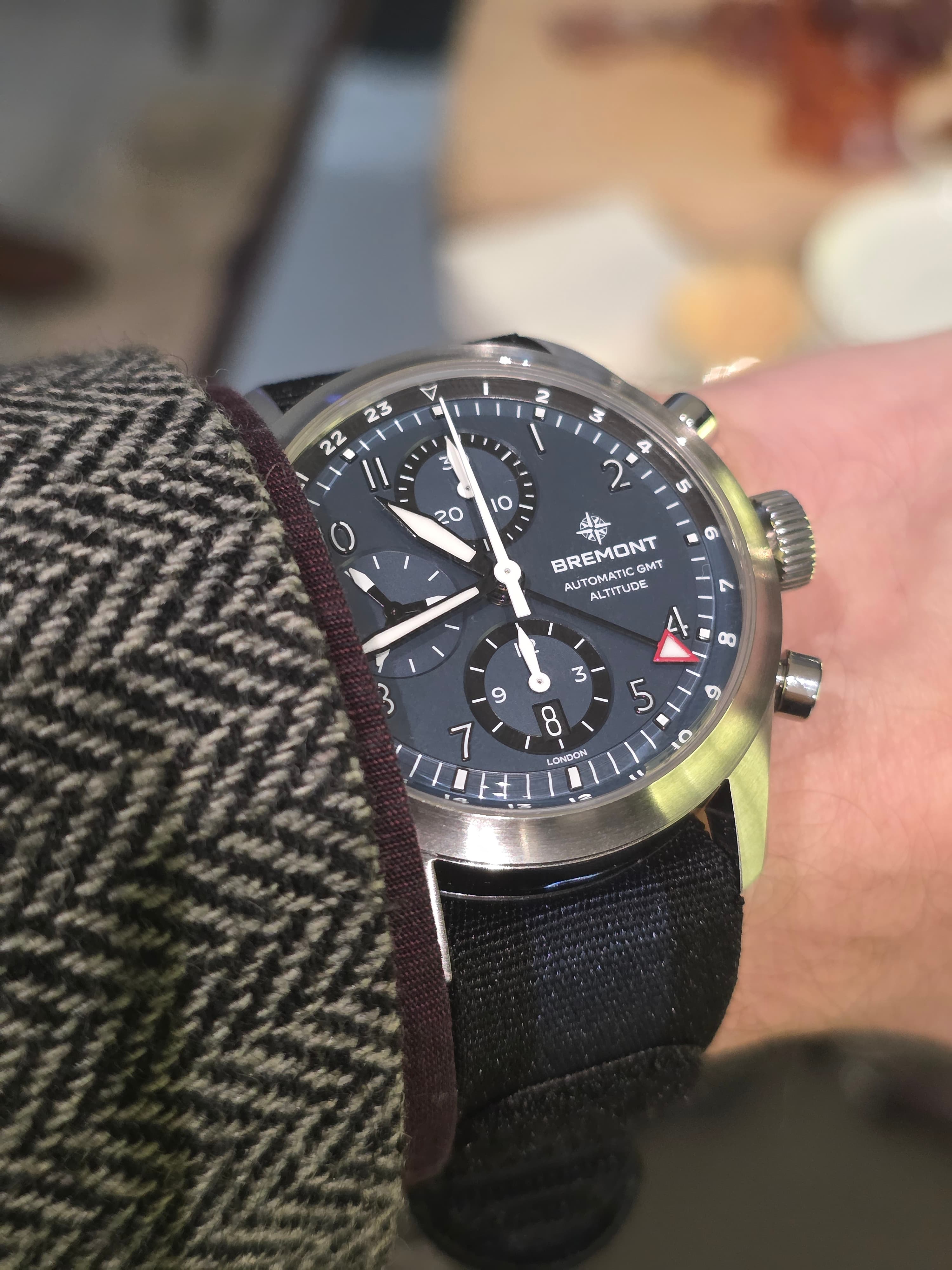 A wristwatch with a black dial and multiple sub-dials, worn on a person's wrist with a herringbone-patterned jacket sleeve.