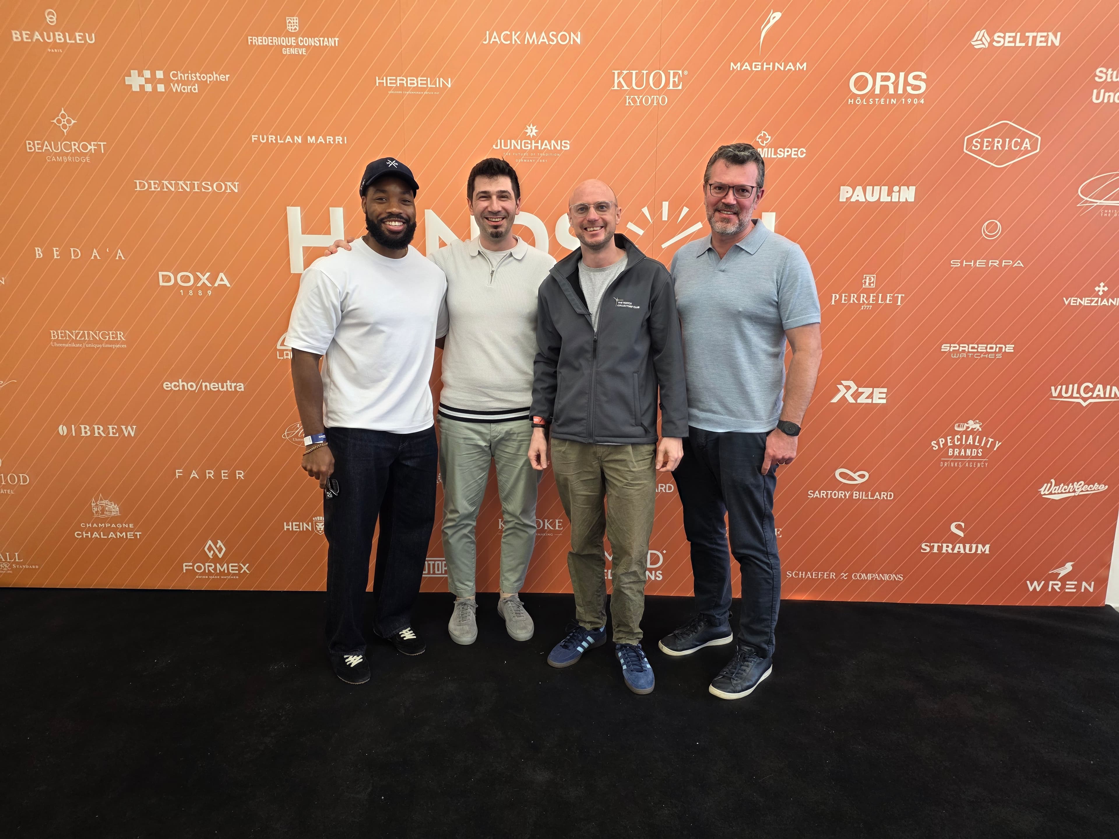 Four watch enthusiasts posing at the entrance to Hands-On Horology Watch show with an orange background and black floor