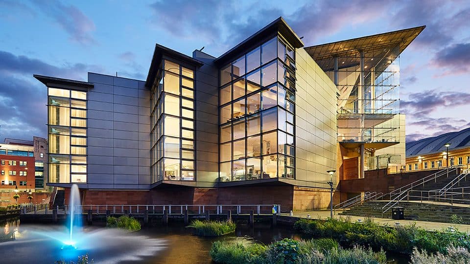 a large angular building with huge windows and silver panelling, the bridgewater hall at dusk with a cloudy blue sky behind it and fountain in front